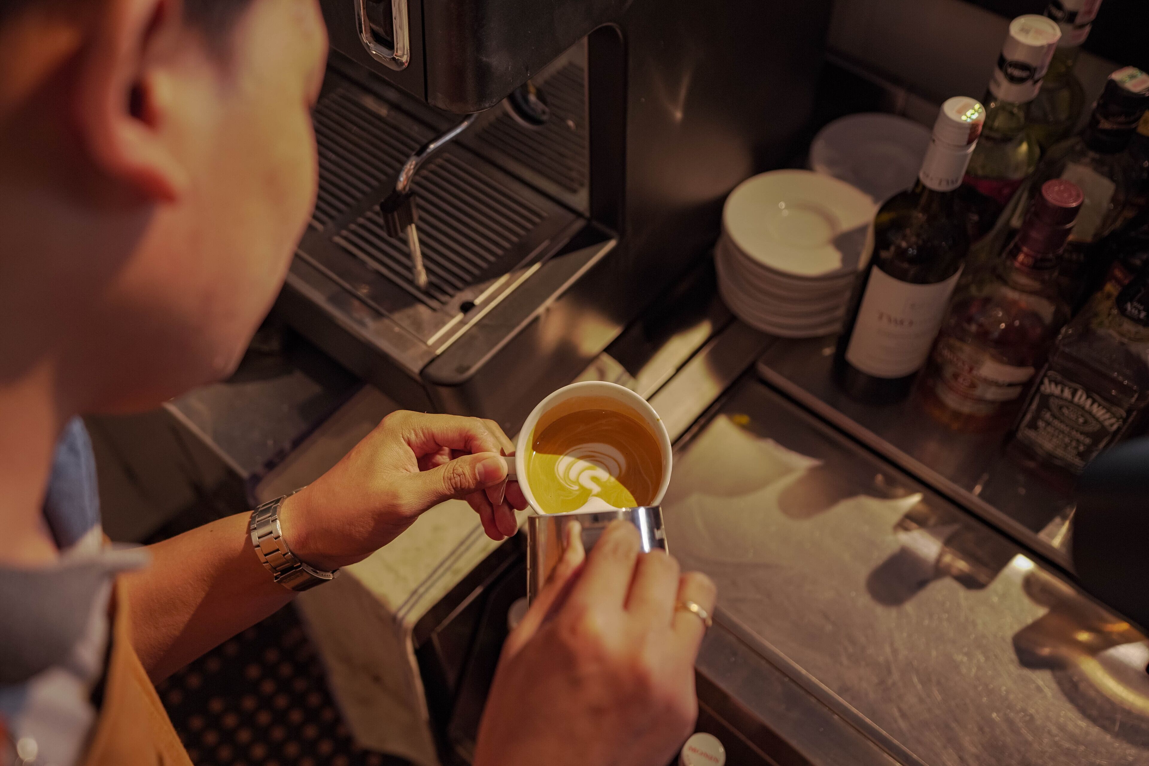 A person holding a cup with a latte art design, a coffee machine, and bottles of alcohol on a counter.