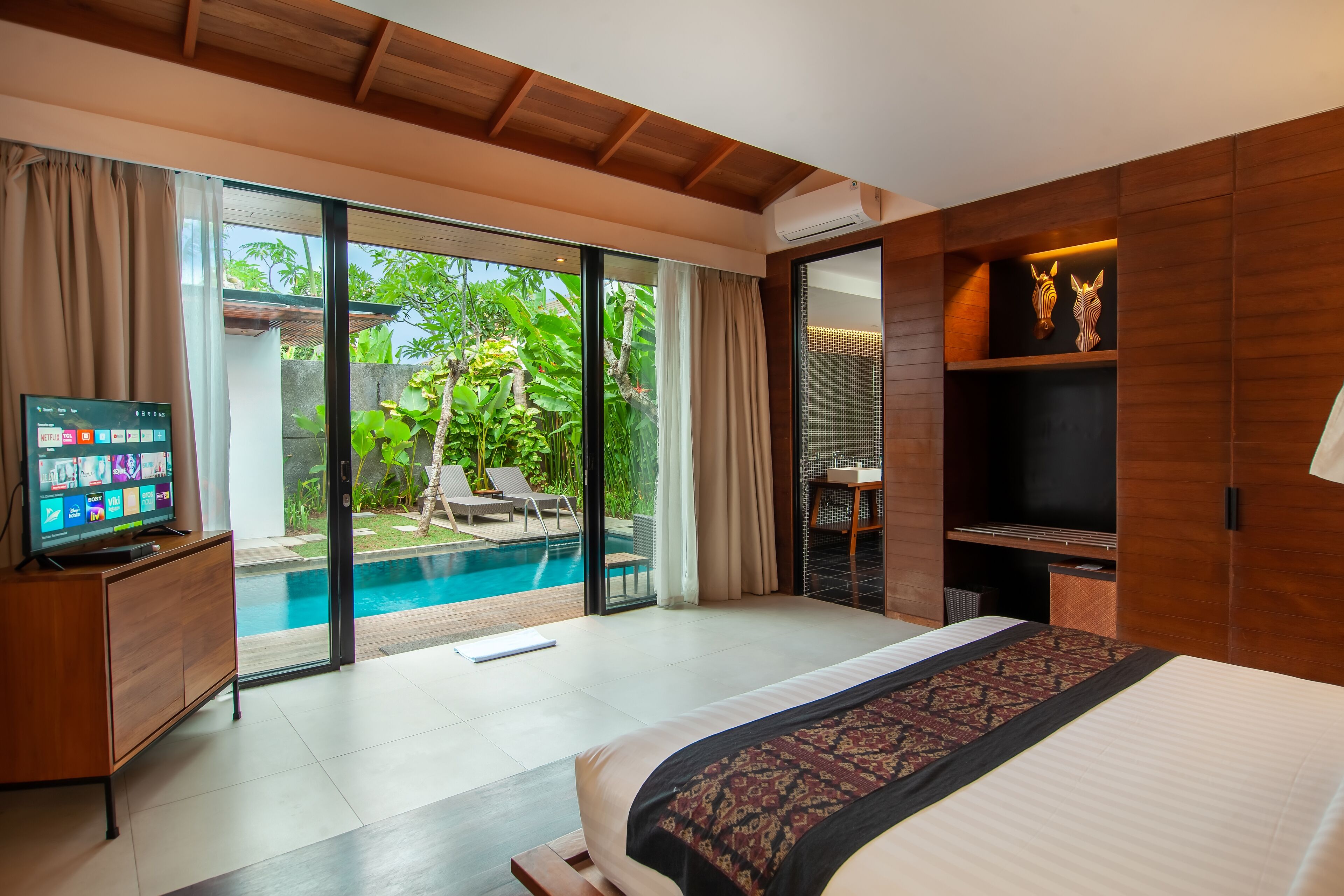 A modern hotel room with a large bed, a TV, and a sliding glass door leading to a pool area.