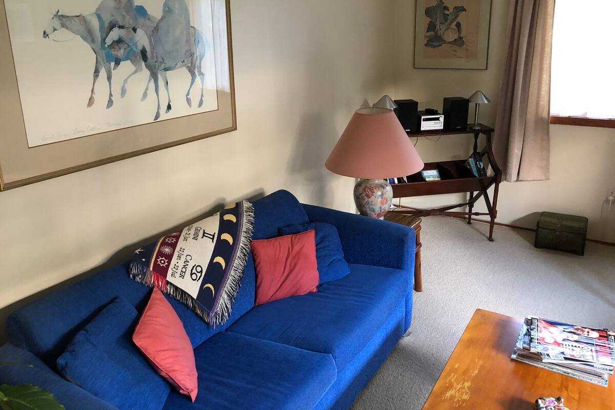 Una sala de estar con un sofá azul, una lámpara, un cuadro enmarcado y una mesa con revistas.