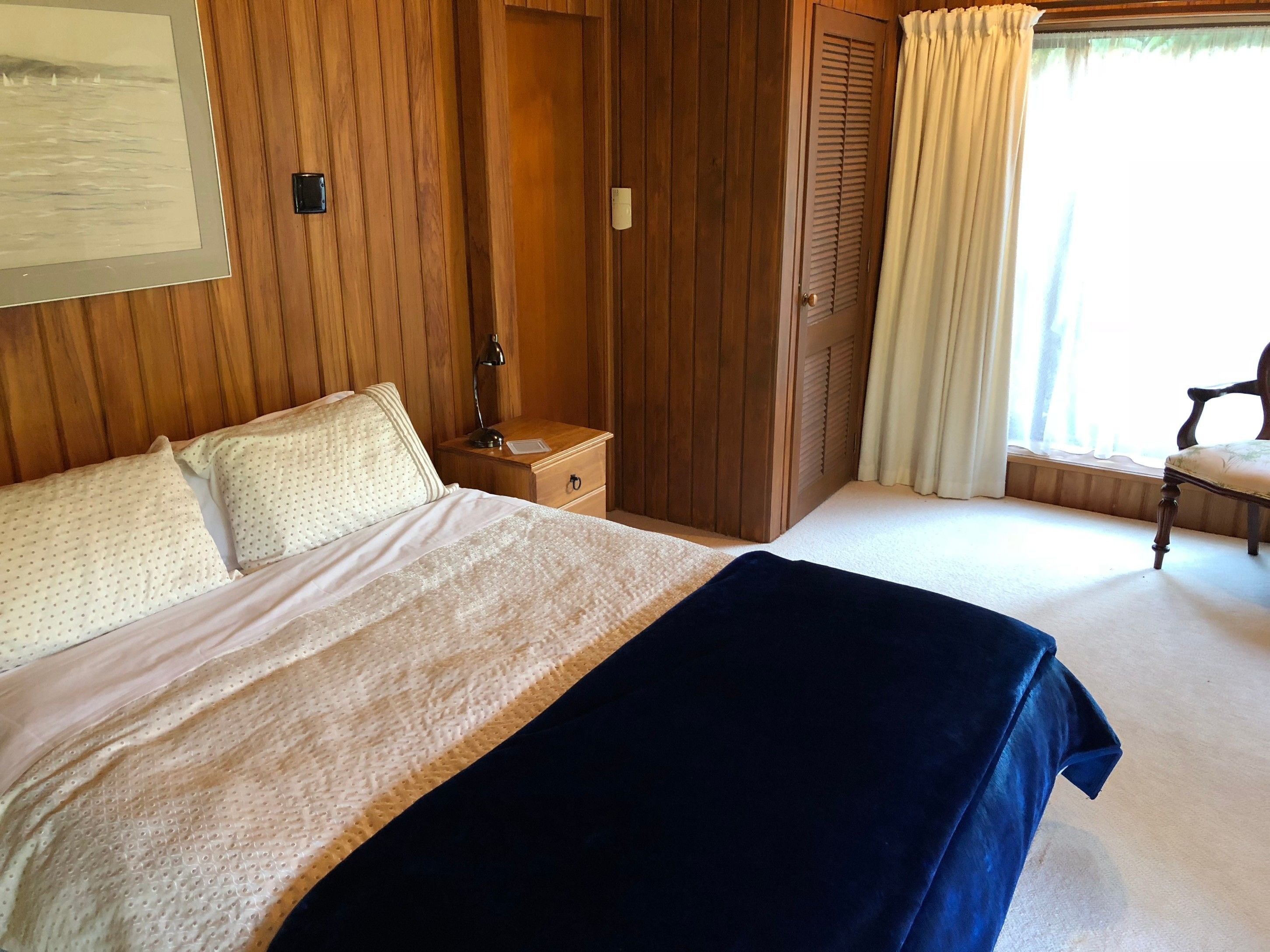 A bedroom with a bed, a nightstand, a wooden wardrobe, a chair, and a window with curtains.