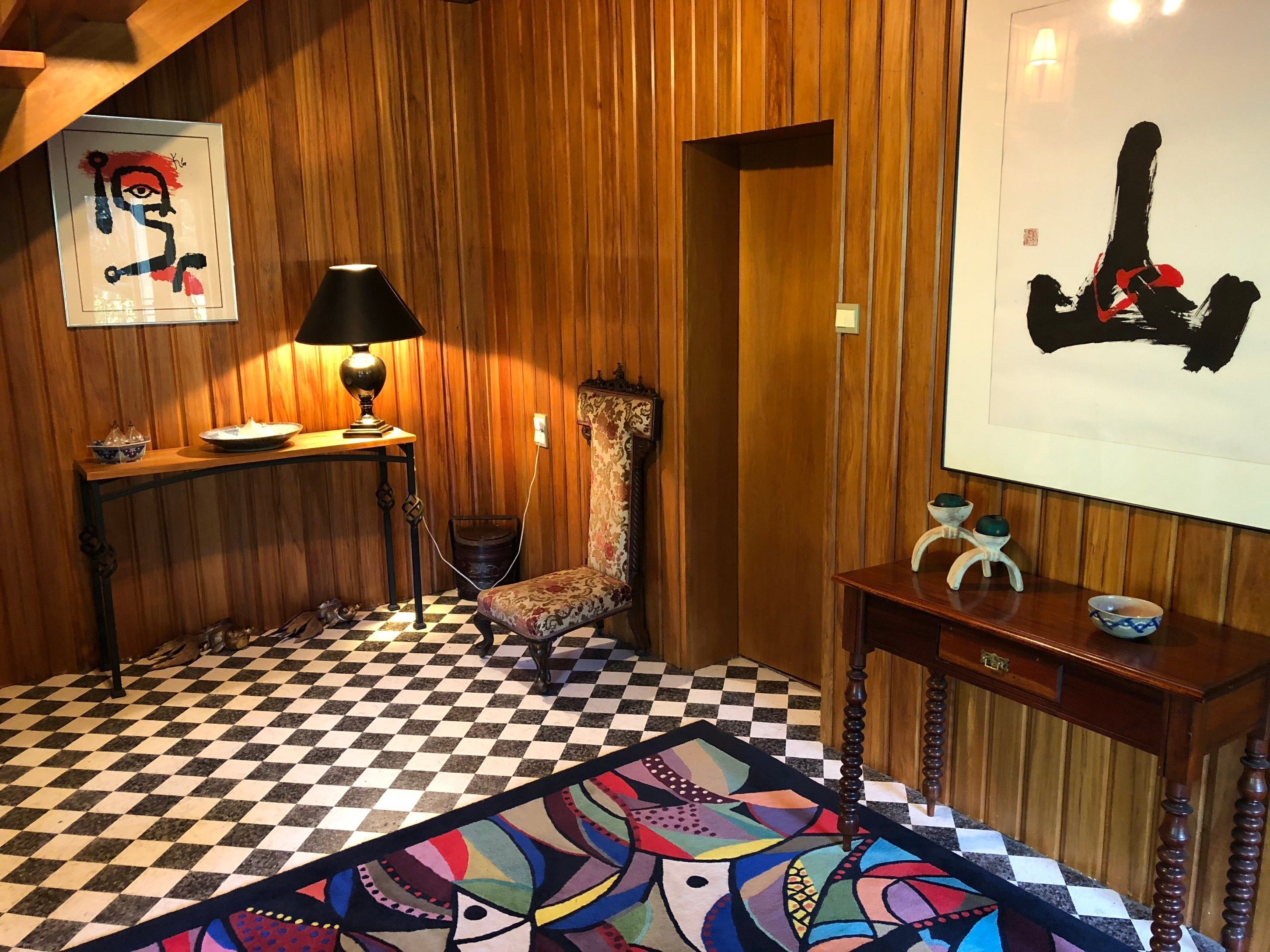 A room with wooden panel walls, a checkered floor, a patterned rug, a lamp, a chair, a table with decorative items, and two framed artworks.