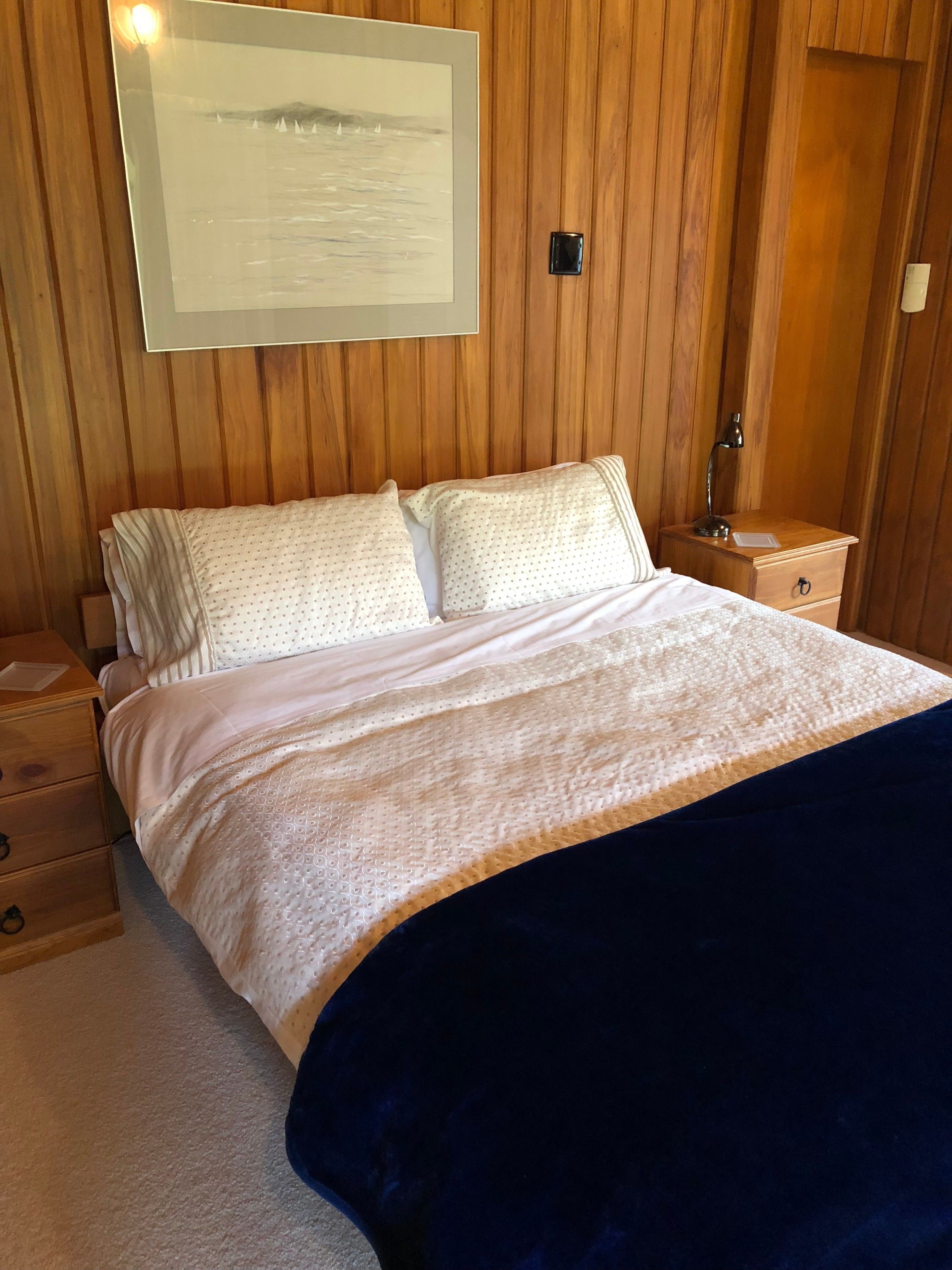 A bedroom with a bed, wooden panel walls, a nightstand, and a framed picture of a landscape.