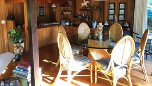 Interior de madera con una mesa redonda de vidrio rodeada de sillas de mimbre, una planta en maceta y varios objetos decorativos en estantes.