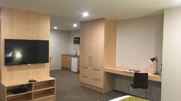 Una habitación de hotel compacta con cama, escritorio, silla, televisor y kitchenette.