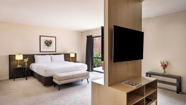 A hotel room with a large bed, bedside tables, a flat-screen TV mounted on a wooden panel, a small bench, and a desk with a lamp and flowers. - Edit Auckland Greenlane (Auckland)