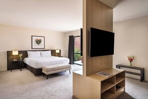 Et hotelværelse med en stor seng, sengeborde, et fladskærms-tv monteret på en træpanel, en lille bænk og et skrivebord med en lampe og blomster.