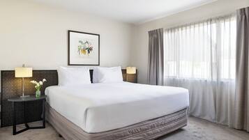 A neatly made bed with white linens, a framed botanical print on the wall, and a window with sheer curtains.
