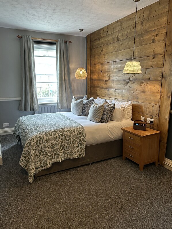 A bedroom with a large bed, a wooden headboard, a bedside table with a radio, and a window with curtains. - The Pelican Inn (Marlborough)