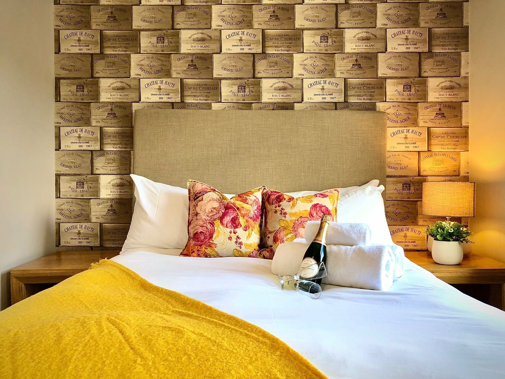 A bed with a floral pillow, a bottle of champagne, and a small plant on a bedside table.