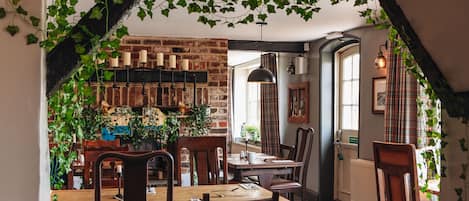 A cozy dining area with brick walls, wooden furniture, and hanging plants.