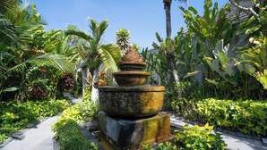 Ein mehrstufiger Steinkaskadenbrunnen, umgeben von üppigen tropischen Pflanzen und einem gepflasterten Weg.