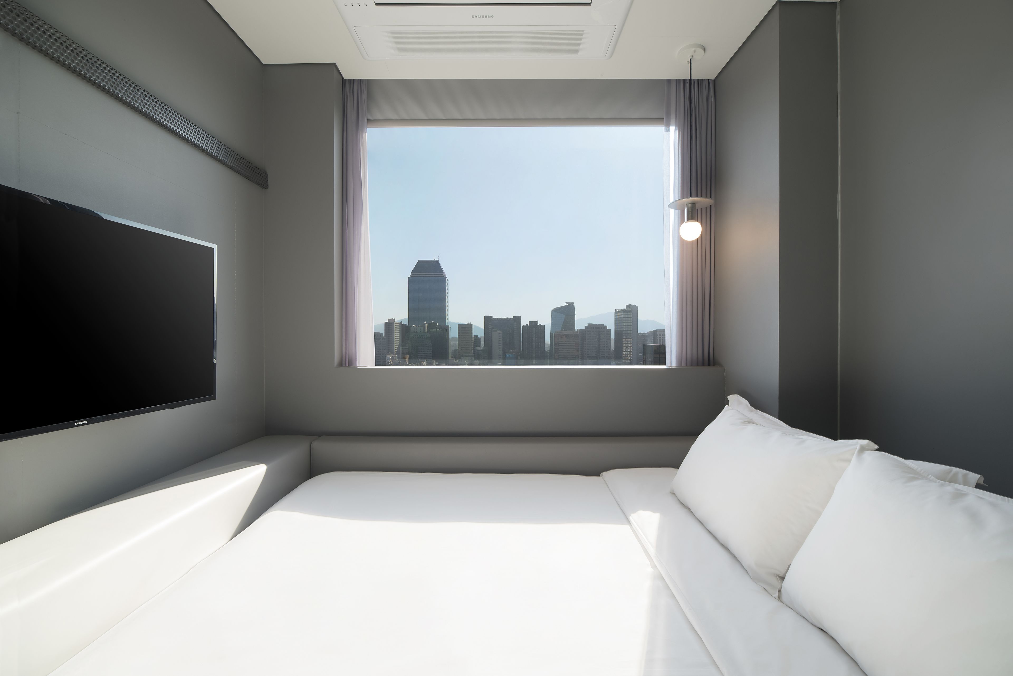 Una camera d'hotel con un'ampia finestra che offre vista sullo skyline cittadino, una televisione a schermo piatto montata a parete e un letto con biancheria bianca.