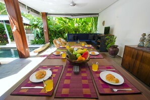 A dining area with a pool view, a fruit bowl, and a remote control on the table. - Pulau Boutique Villas (Kerobokan)