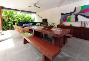 A modern living room with a wooden dining table, bench, and blue sofa with green cushions. There is a large abstract painting on the wall and a ceiling fan.
