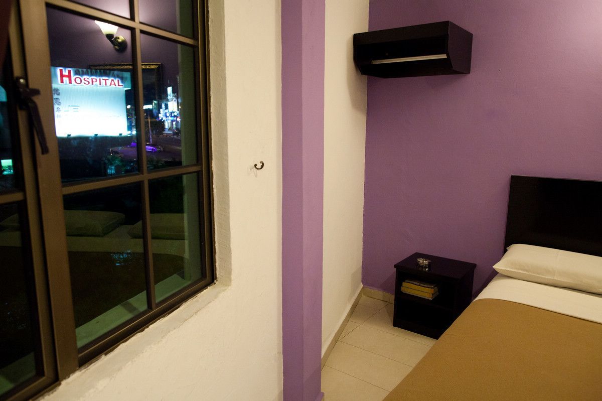 A hotel room with a bed, a nightstand, a window with a view of a hospital sign, and a purple wall.