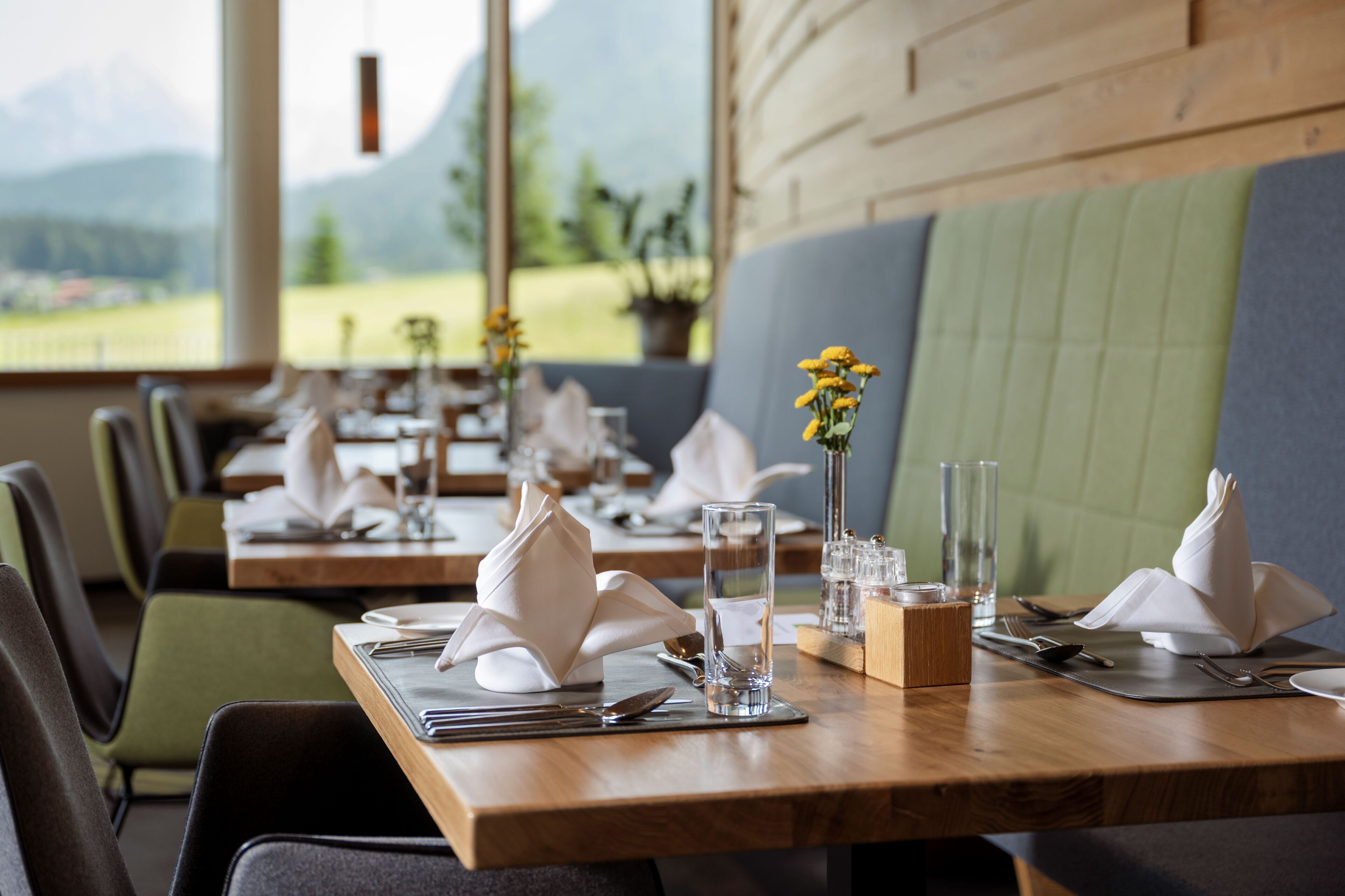 A restaurant with tables set for dining, a view of mountains, and a green cushioned bench.
