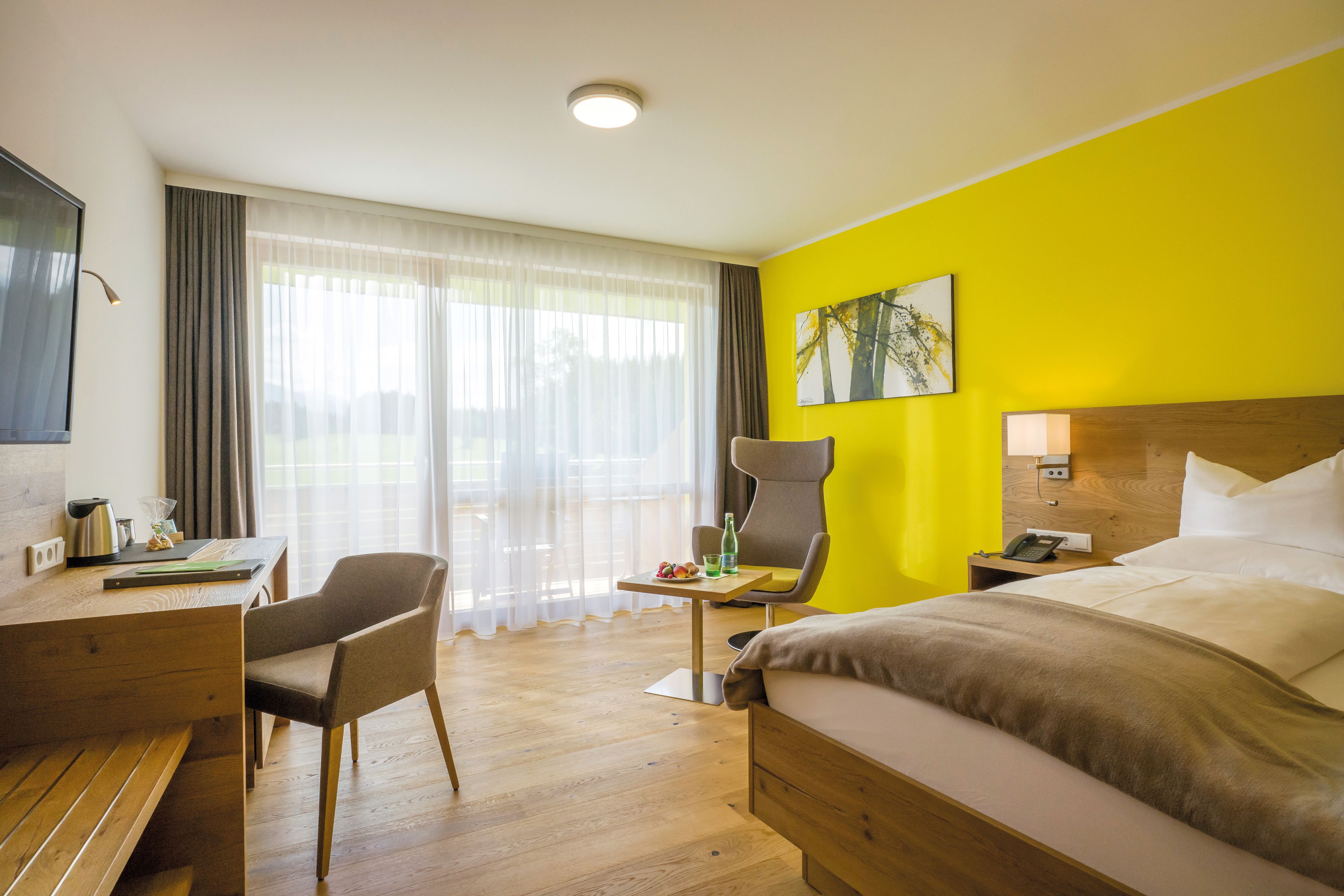 A hotel room with a bed, a desk, a chair, a table with a bottle and fruit, and a window with curtains.