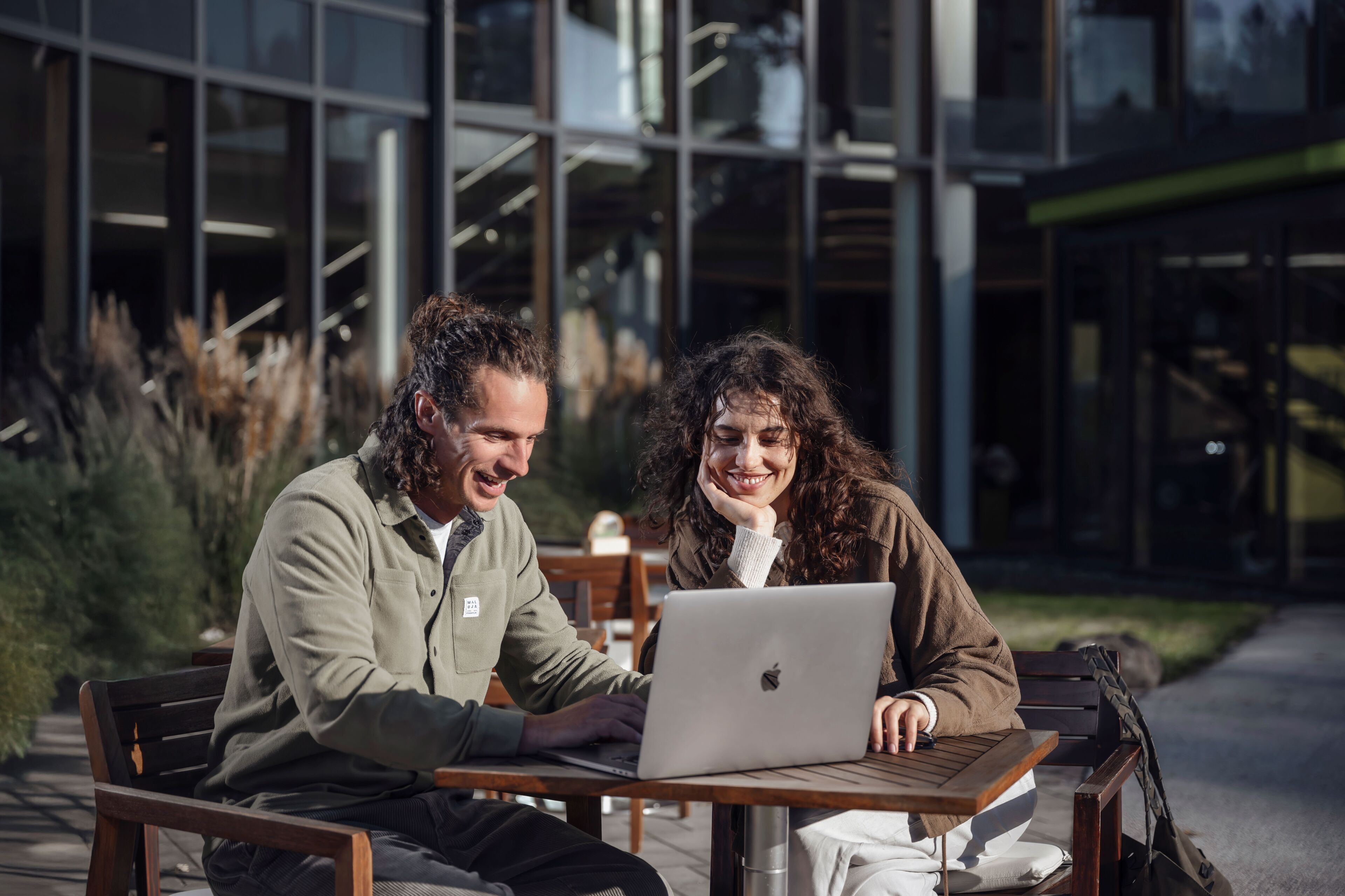 Dos personas sentadas en una mesa al aire libre con una computadora portátil, frente a un edificio moderno con grandes ventanales.