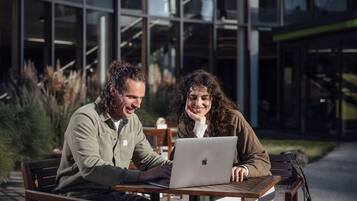 Dos personas sentadas en una mesa al aire libre con una computadora portátil, frente a un edificio moderno con grandes ventanales.