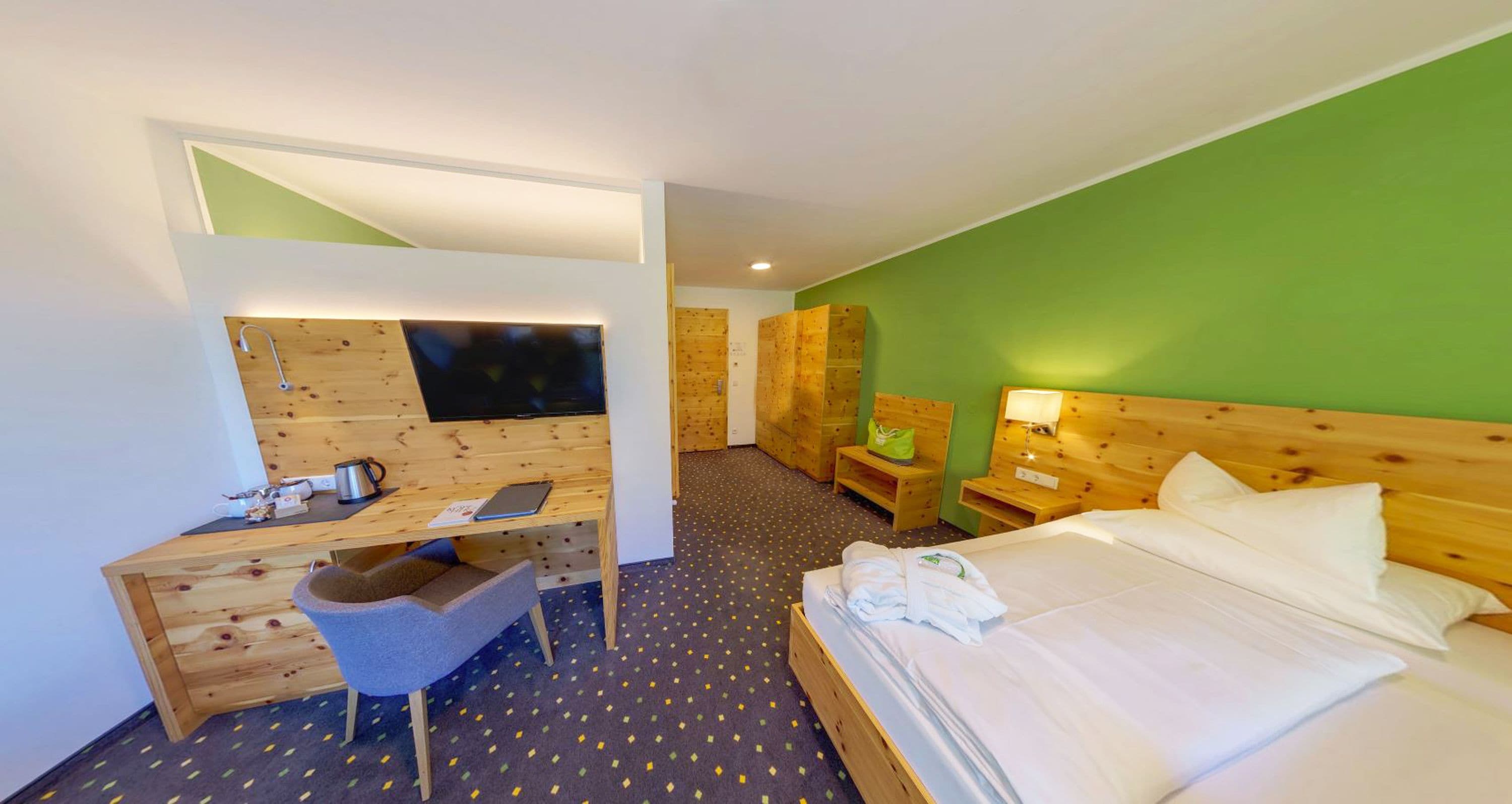 A hotel room with a wooden desk, a chair, a bed with white linens, and a green accent wall.