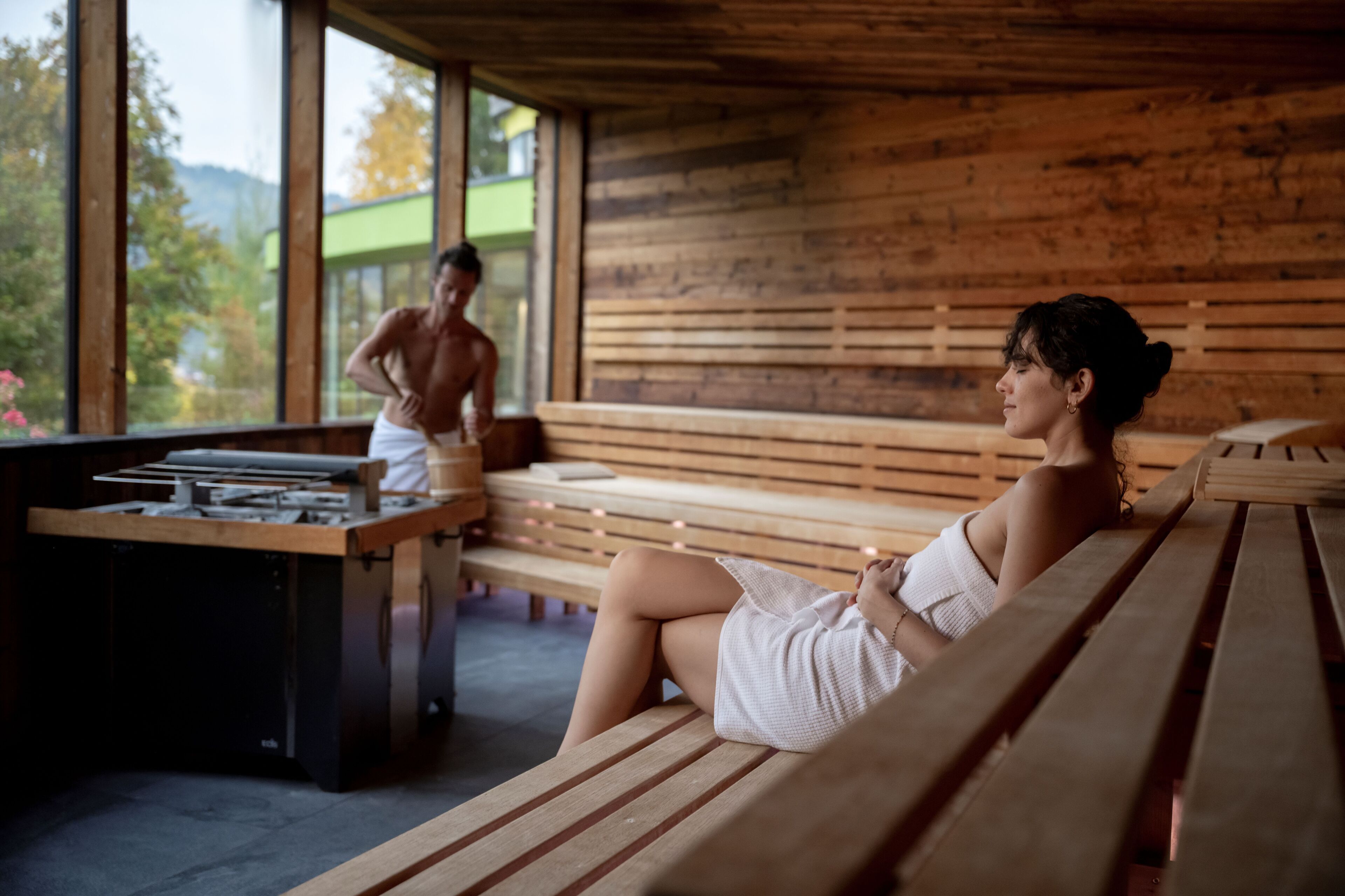 A wooden interior with benches and a table, large windows, and a view of greenery outside.