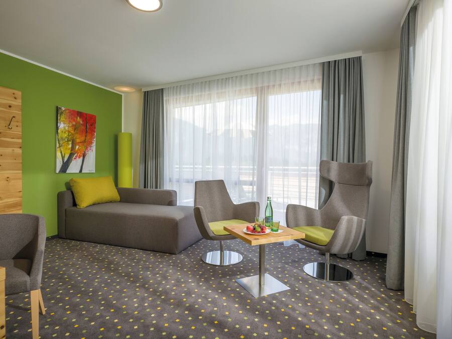 A modern hotel room with a green accent wall, a large window with curtains, a sofa, two armchairs, a small table with a bottle and glass, and a patterned carpet.