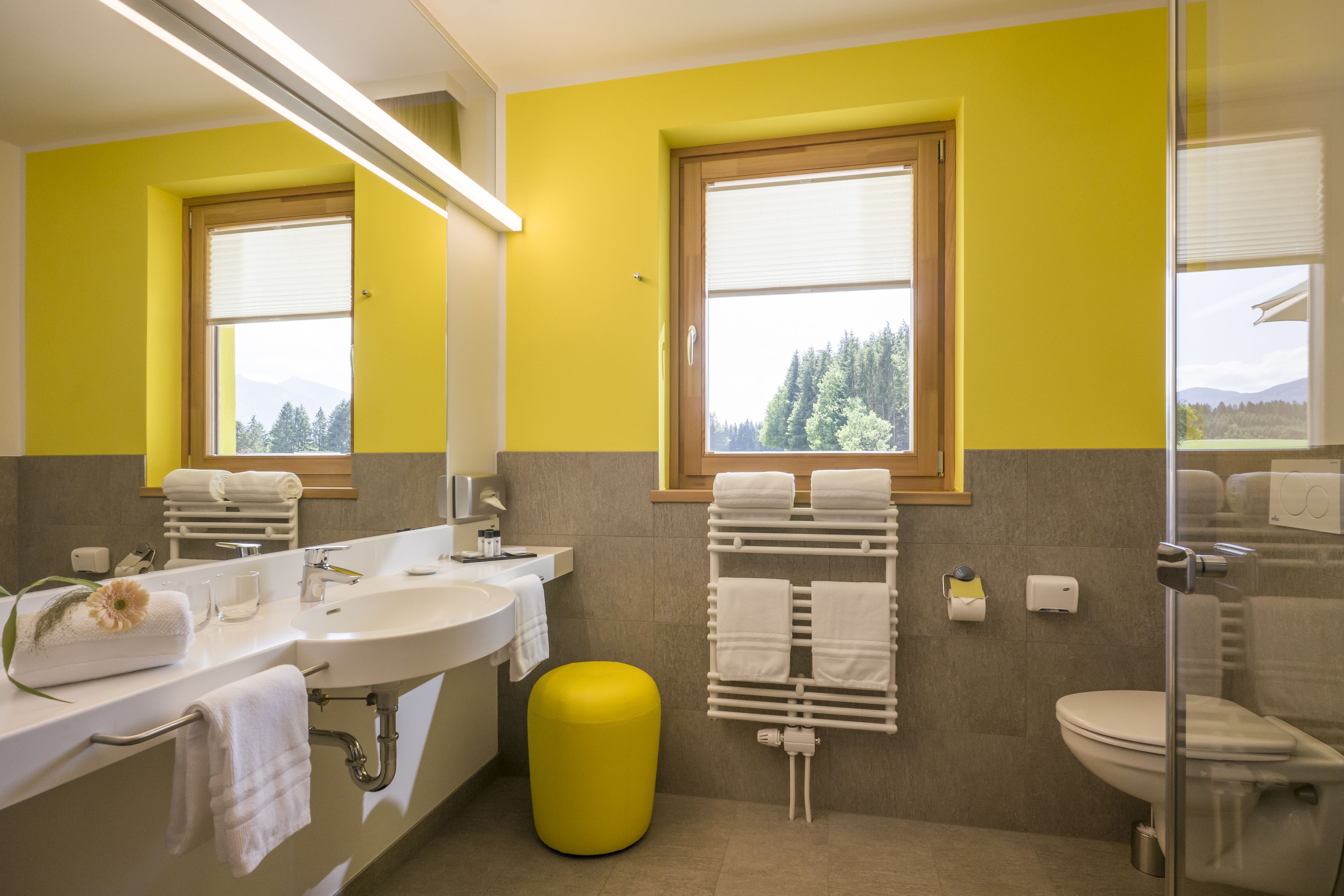 Un baño con una pared amarilla, un ventanal grande, un lavamanos, un inodoro y un taburete amarillo.