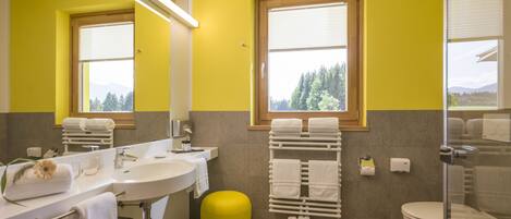 Un baño con una pared amarilla, un ventanal grande, un lavamanos, un inodoro y un taburete amarillo.
