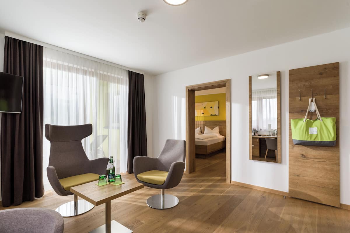A modern hotel room with a wooden floor, a sofa, two armchairs, a small table with a bottle and glasses, a bed with a yellow headboard, a mirror, and a green bag hanging on the wall.
