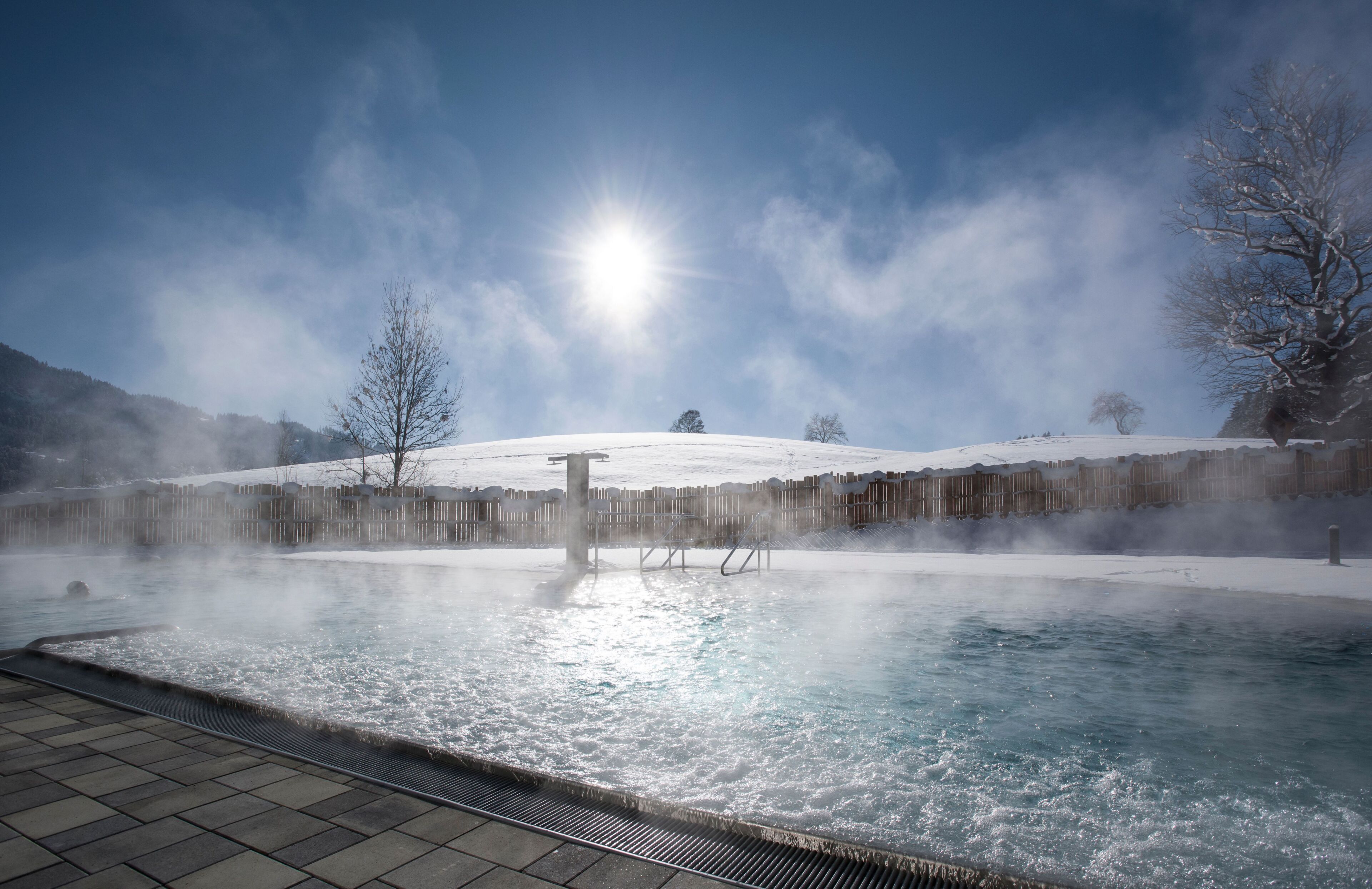 A hot spring pool with steam rising, surrounded by snow-covered hills and a clear sky.