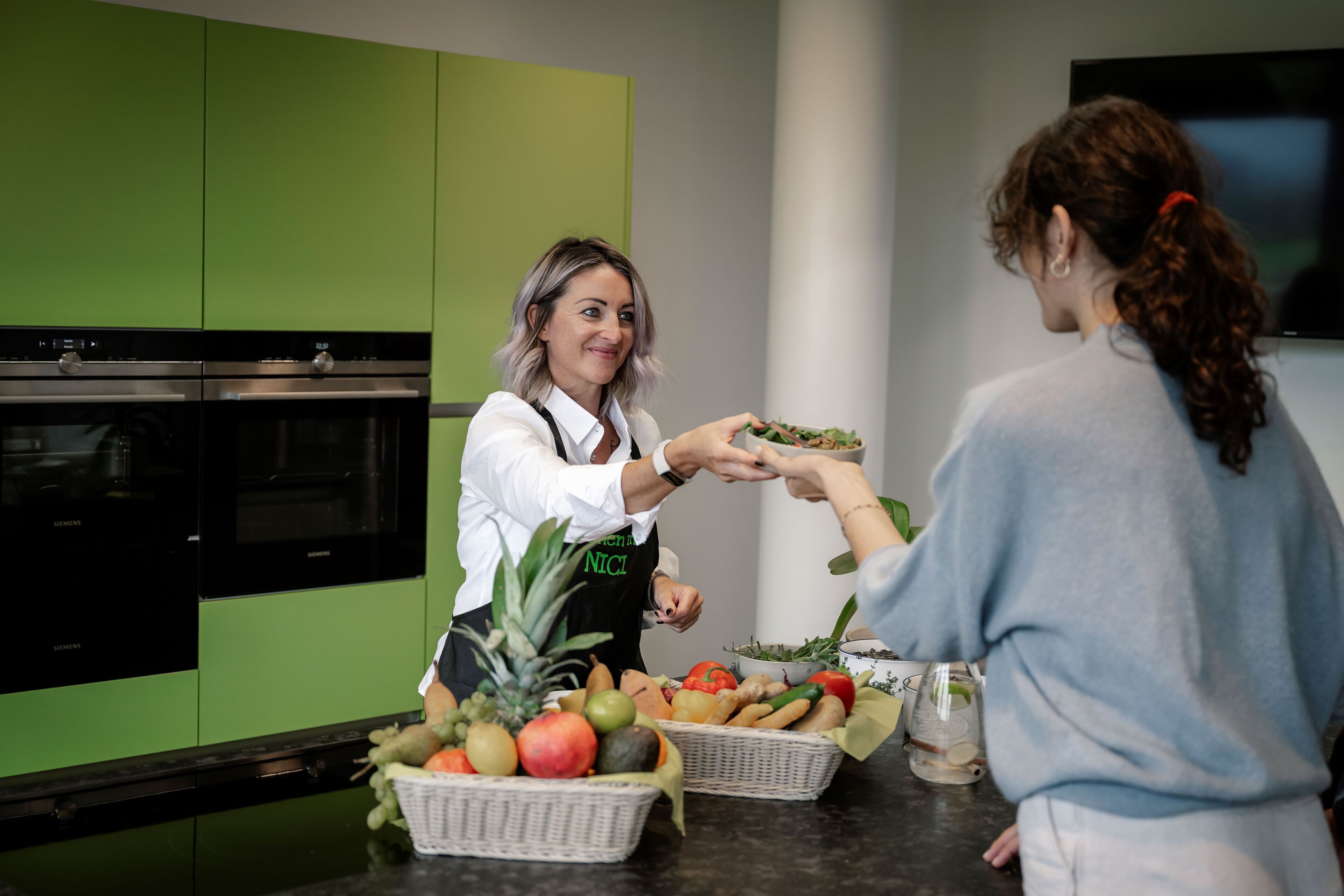 A kitchen with green cabinets, a stainless steel oven, and a person handing a dish to another person.