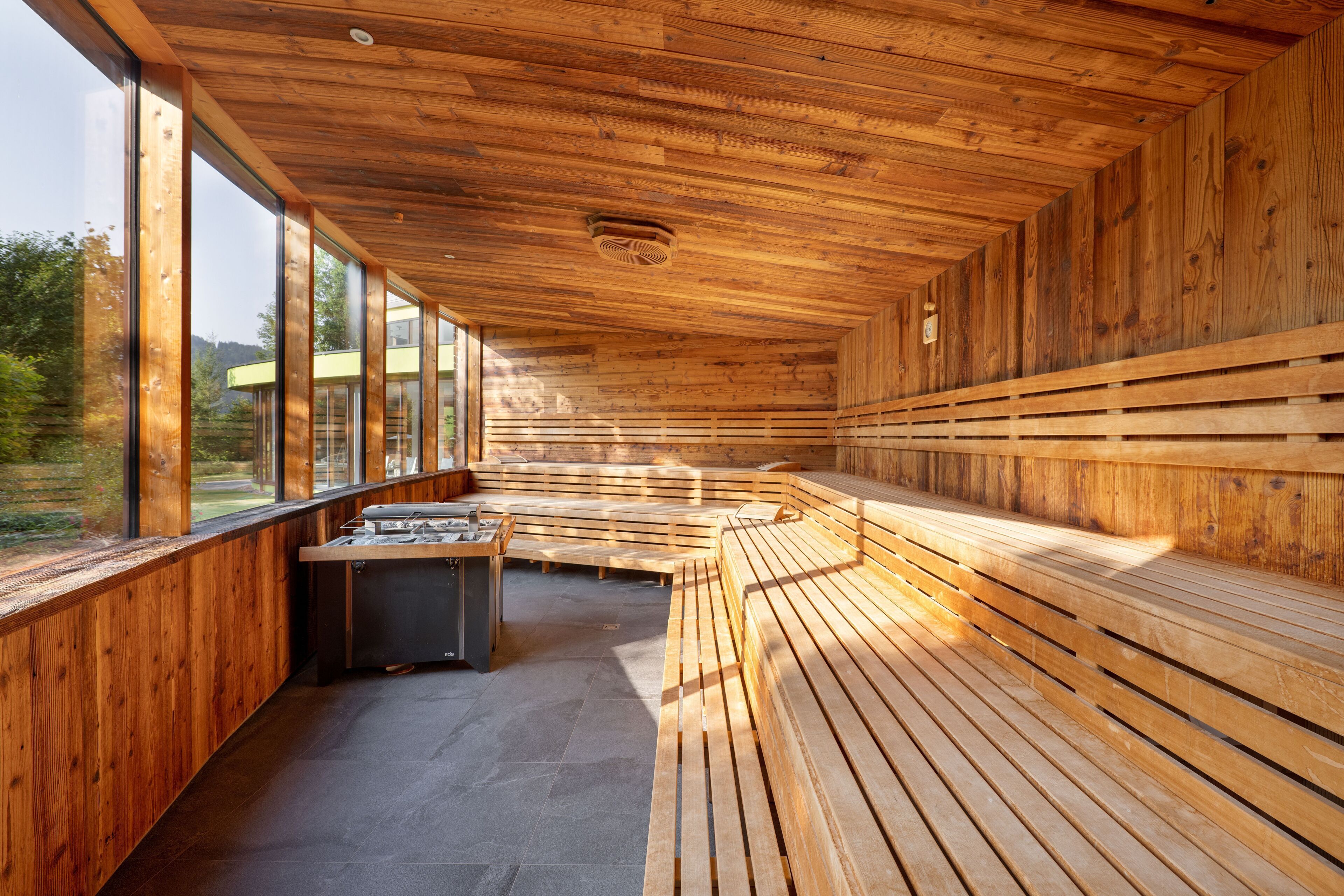 A sauna with wooden benches and a stone floor, large windows, and a wooden ceiling.
