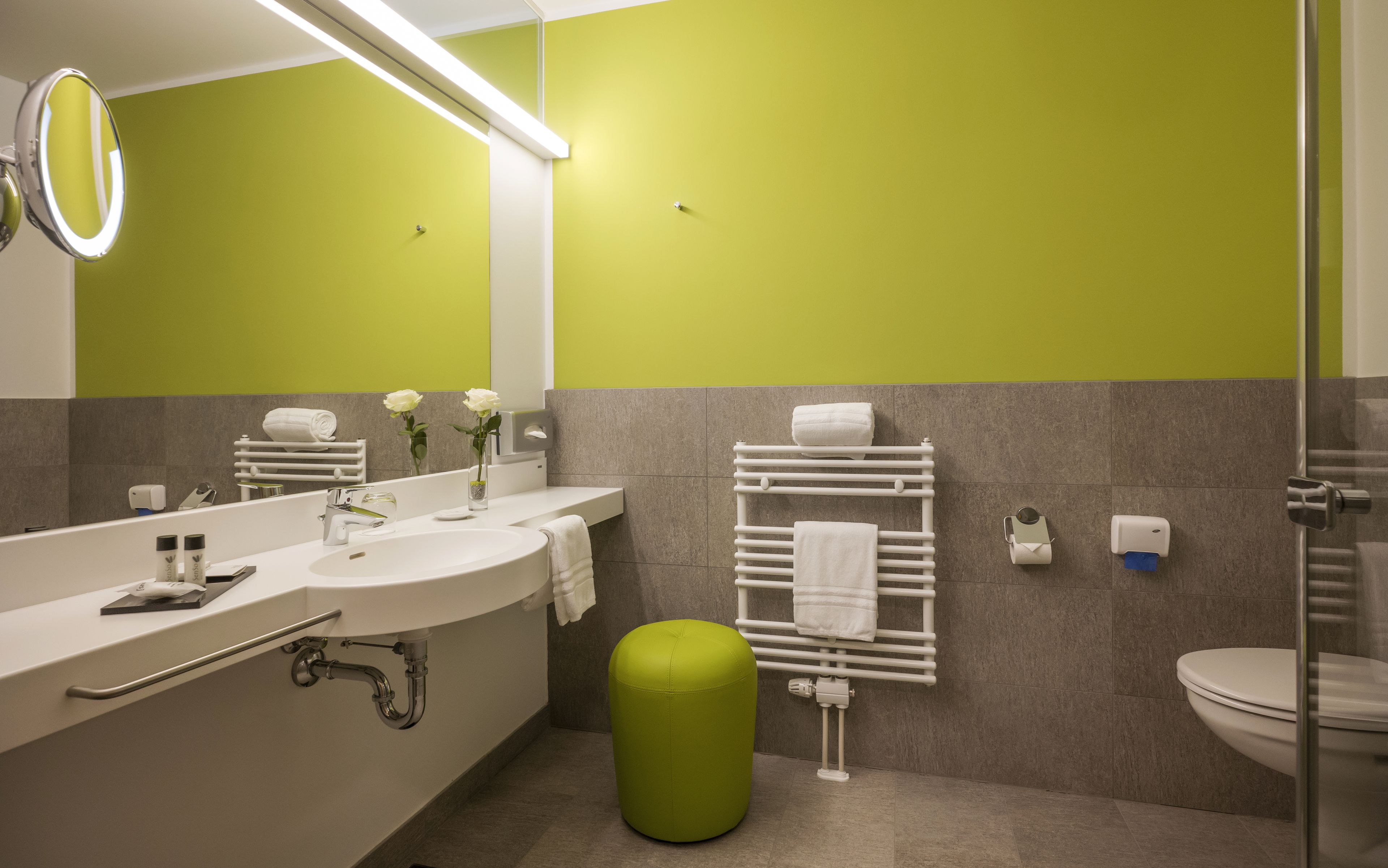 A modern bathroom with a green accent wall, a white sink, a mirror, a toilet, and a green ottoman.