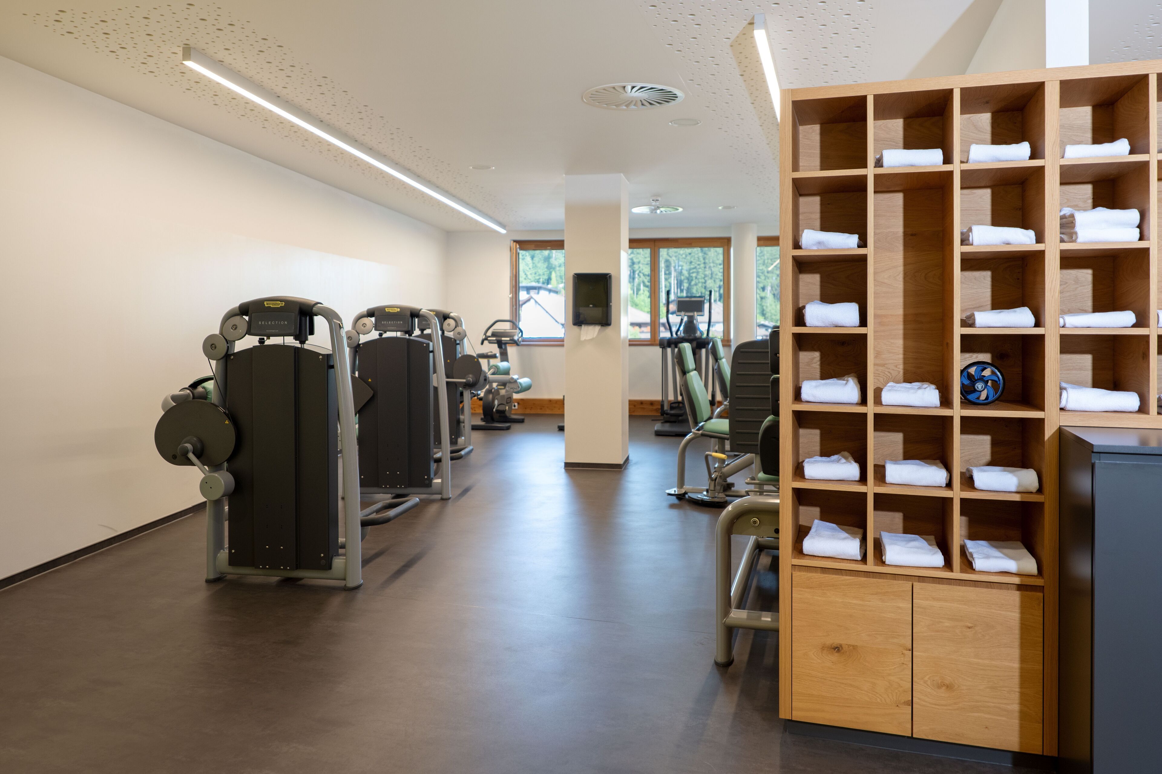 A gym with exercise equipment, a wooden towel rack, and a view of trees through large windows.