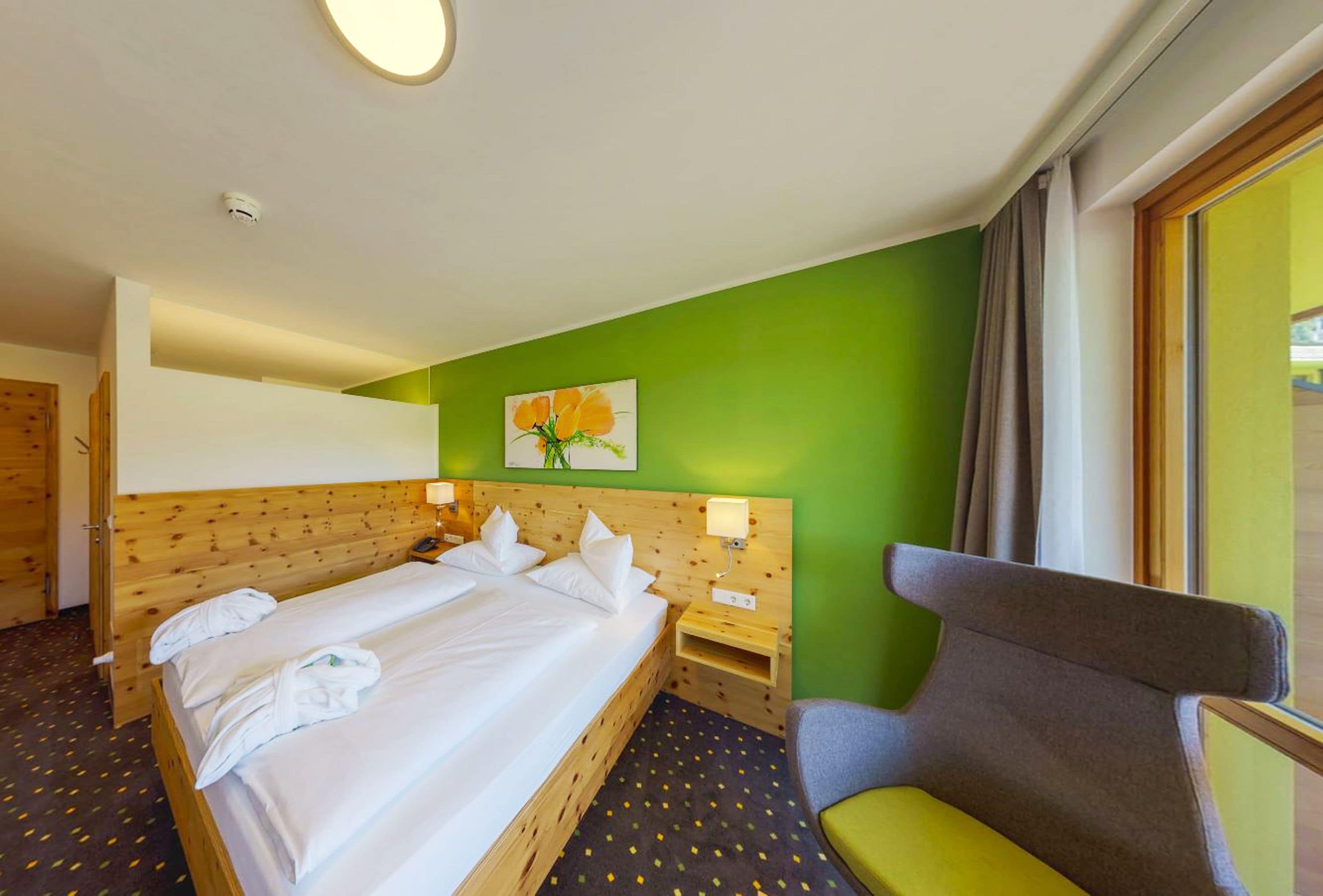 Ein Hotelzimmer mit einem hölzernen Kopfteil, einem Bett mit weißer Bettwäsche, einem Nachttisch mit Lampe, einer grünen Wand und einem Fenster mit Vorhängen.