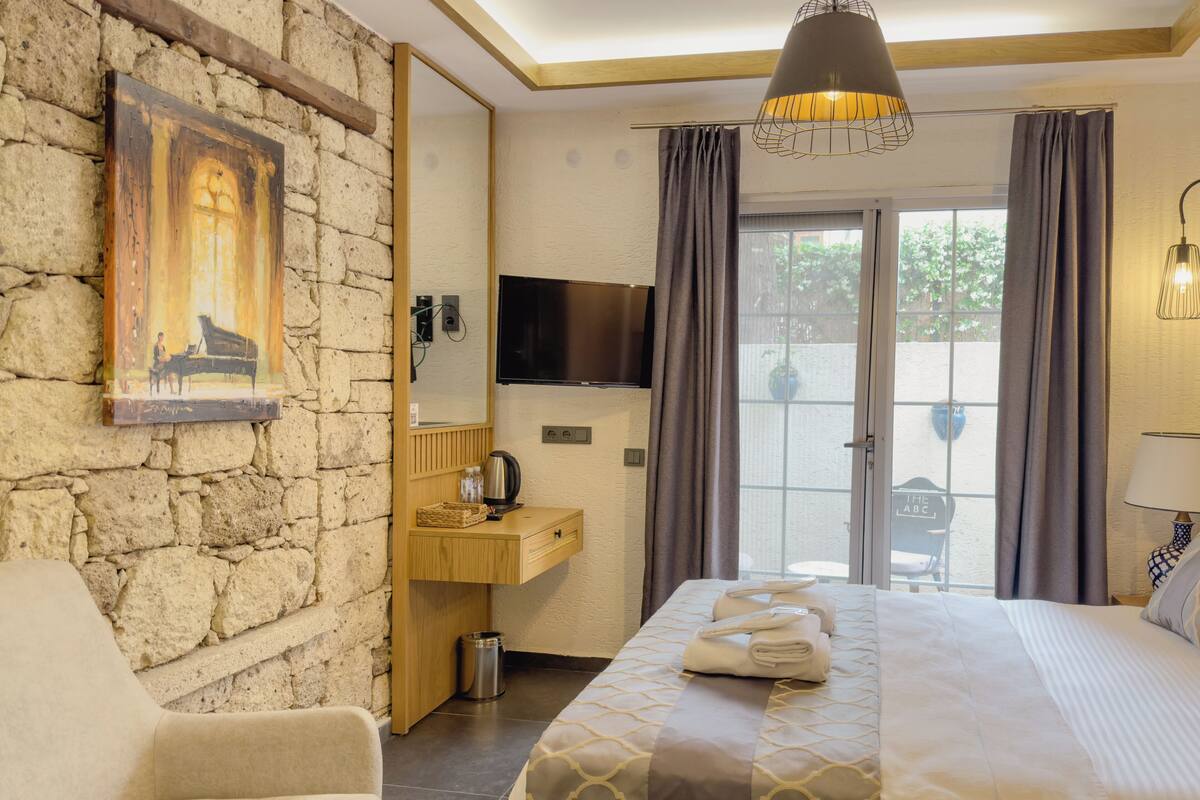 A hotel room with a stone wall, a painting, a bed with a patterned bedspread, a bedside table, a TV, and a view of the outdoors.