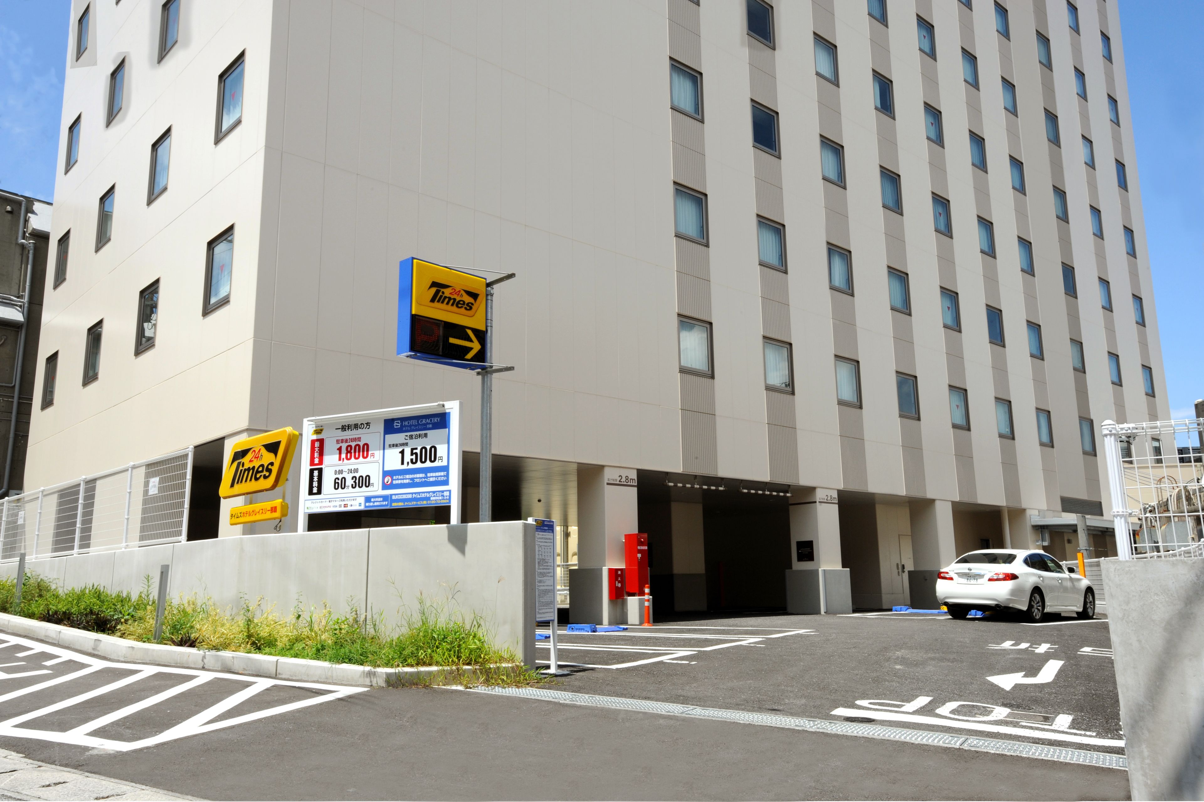 A hotel entrance with a Times sign, a parking area, and a white car parked in front.