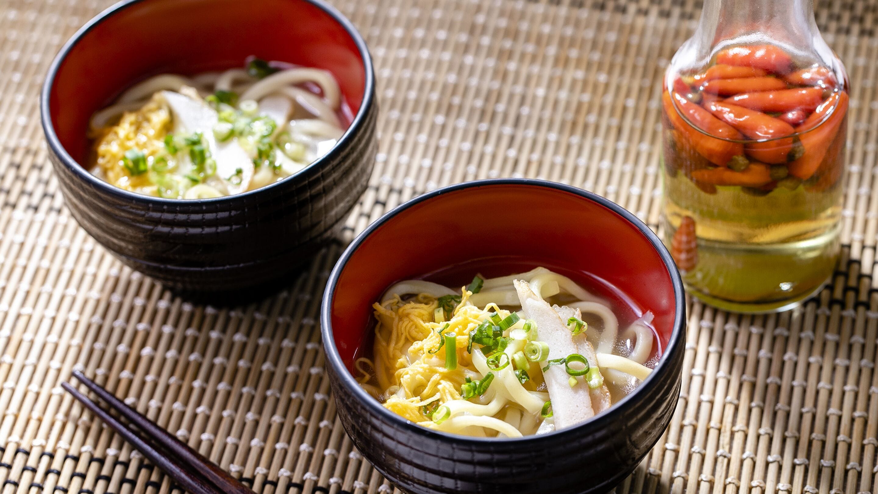 Two bowls of ramen with toppings, a bottle of soy sauce, and a jar of pickled vegetables on a bamboo mat.