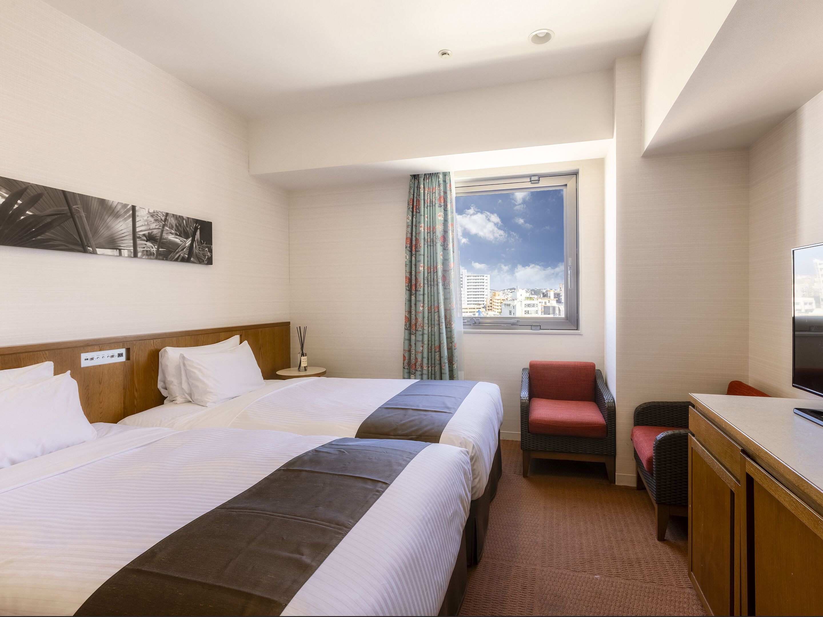 A hotel room with two beds, a desk with a television, a chair, a window with a city view, and a small table with a lamp.