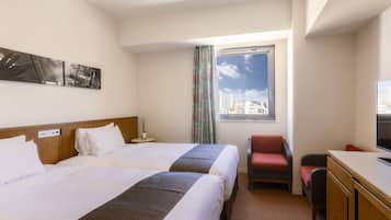 A hotel room with two beds, a desk with a television, a chair, a window with a city view, and a small table with a lamp.