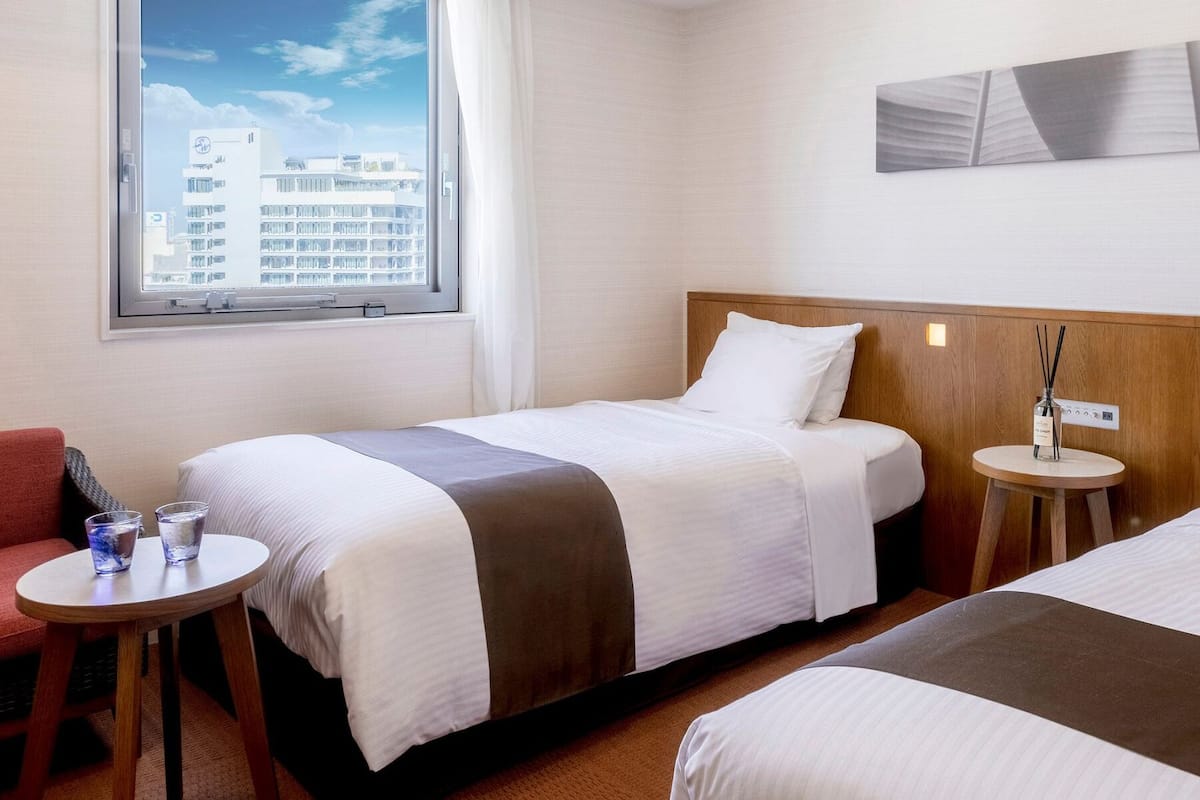 Hotellihuone, jossa on kaksi sänkyä, pieni pöytä, tuoli ja ikkuna, josta on näkymä kaupunkiin.