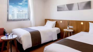 Habitación de hotel con dos camas, una mesita, una silla y una ventana con vista a la ciudad.