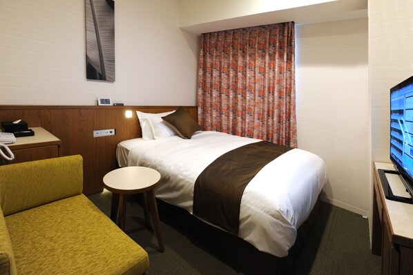 Une chambre d’hôtel avec un lit, une petite table ronde, un fauteuil jaune, une télévision et un téléphone posés sur une table de chevet en bois.