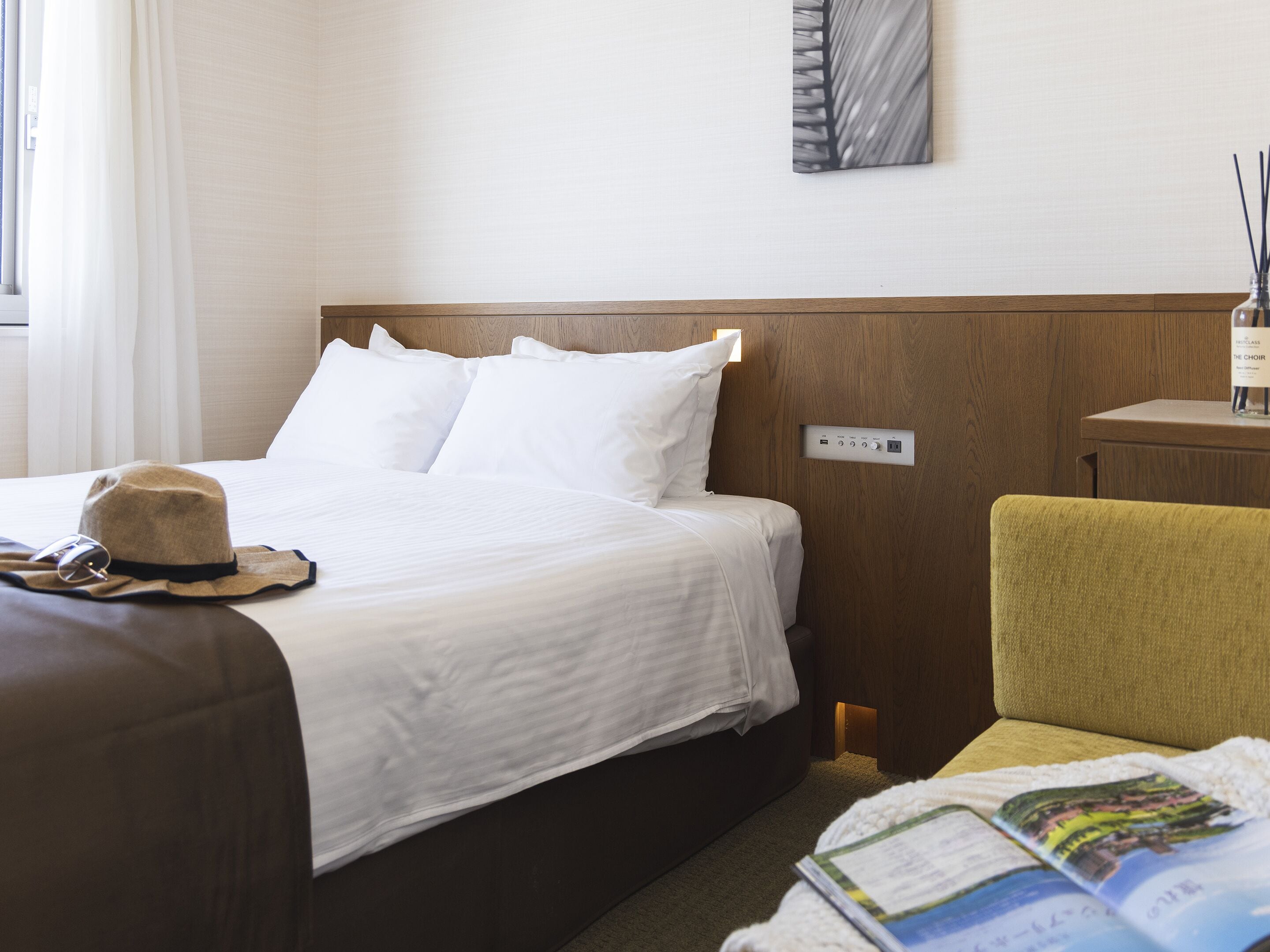 A hotel room with a bed, a bedside table with a lamp, a chair, and a magazine on the floor.