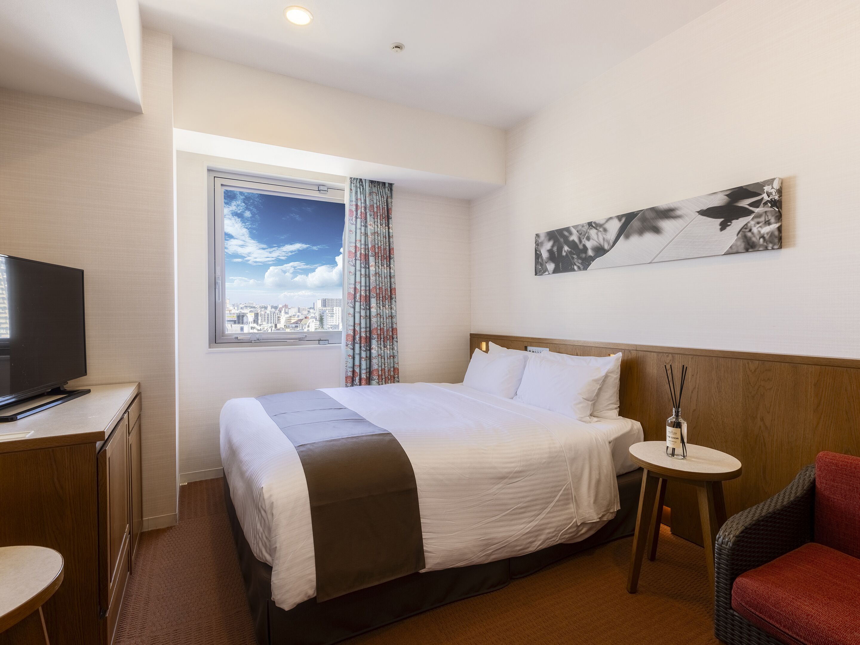 A hotel room with a bed, a television on a desk, a window with a city view, and a small table with a bottle.