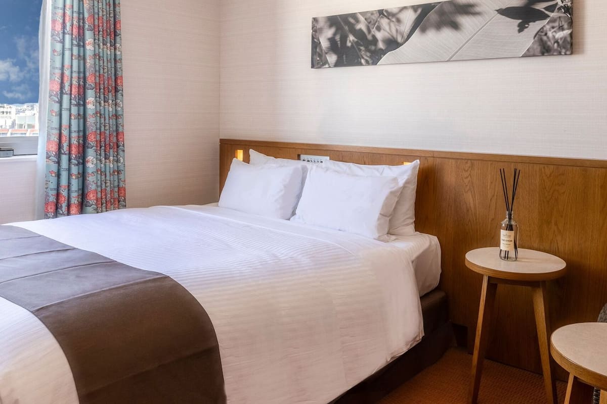 Hotellihuone, jossa on sänky, pieni pyöreä pöytä, punainen tuoli, ikkuna, josta on näkymä kaupunkiin, ja seinälle kiinnitetty taideteos.