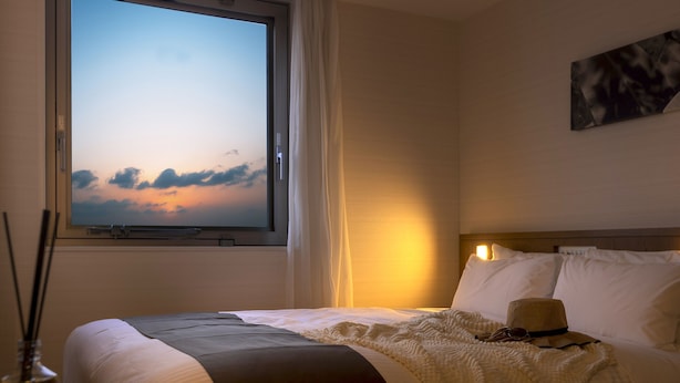Habitación de hotel con cama, ventana con vista al cielo, un cuadro en la pared y una botella en la mesita de noche.