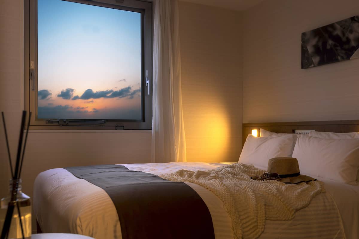 Hotellihuone, jossa on sänky, ikkuna, josta avautuu näkymä taivaalle, taulu seinällä ja pullo yöpöydällä.