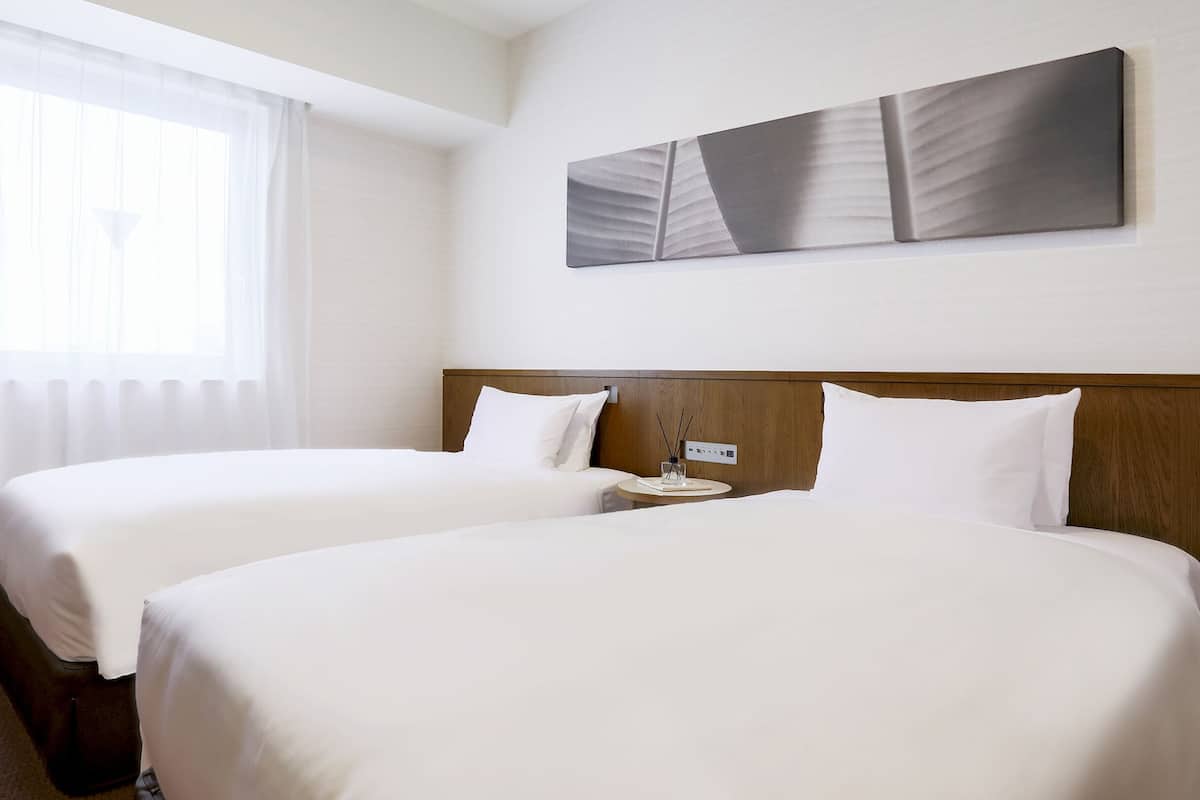 Hotellihuone, jossa on kaksi sänkyä, puinen pääty, yöpöytä lampun kanssa ja ikkuna, jossa on ohuet verhot.