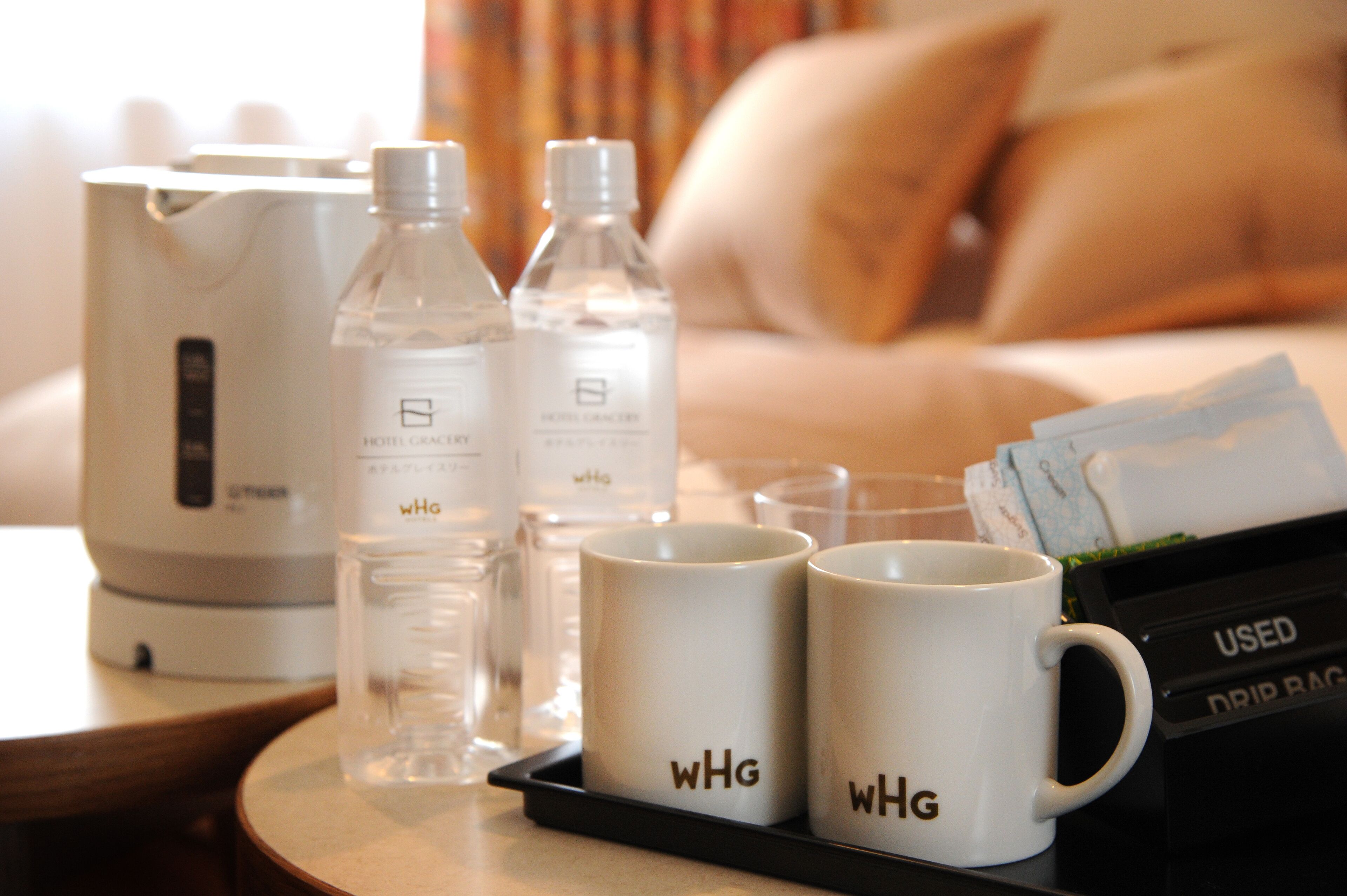 Habitación de hotel con una cama, una mesita de noche con una tetera, dos botellas de agua, dos tazas blancas con la inscripción 